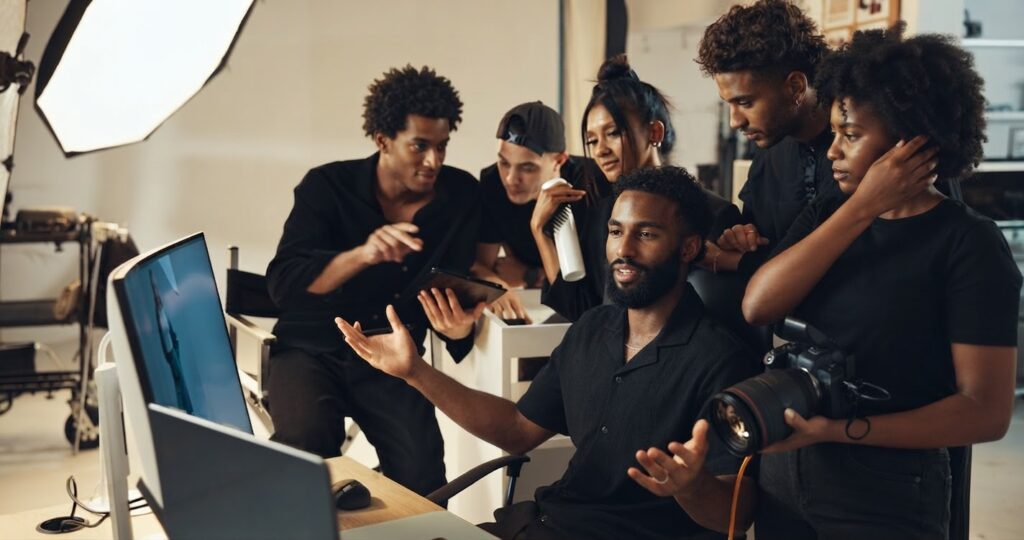 Six people from Rubberband Communications gather in a photography studio around a computer screen, discussing something; one person holds a camera, while the others listen and look at the monitor. - Proudly Developing