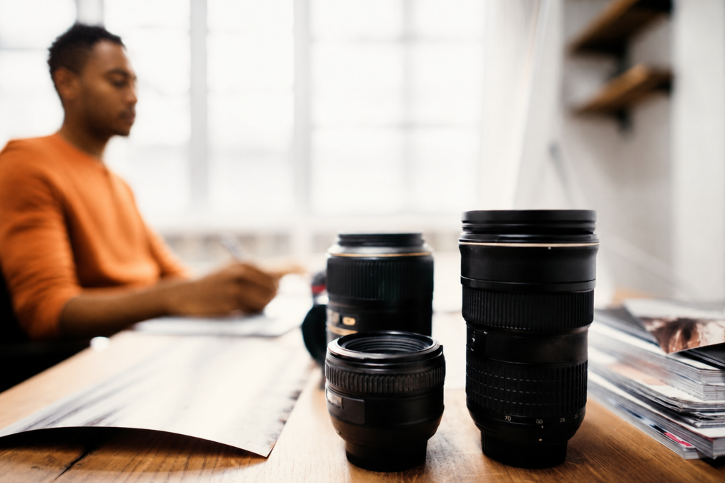 Three camera lenses are arranged on a wooden table in the foreground, with a person in an orange shirt working at a desk in the blurred background—capturing a glimpse into the creative workspace of Rubberband Communications. - Proudly Developing
