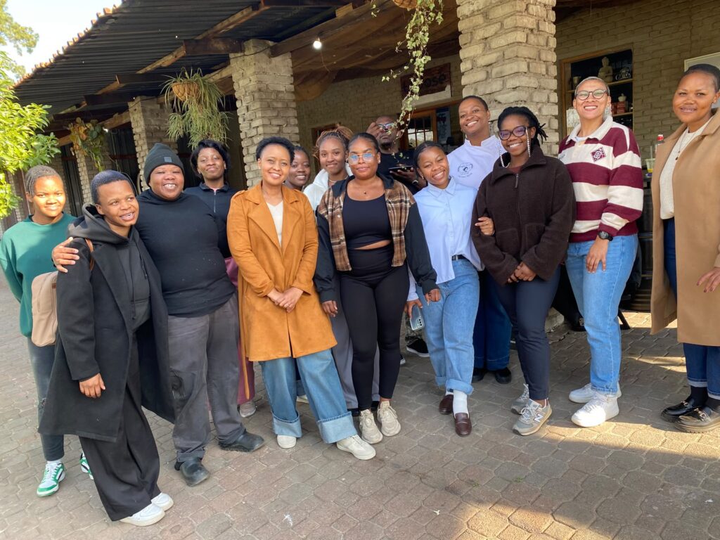 A group of thirteen people stand together outdoors in front of a brick building, smiling at the camera after completing First Aid Training. - Proudly Developing