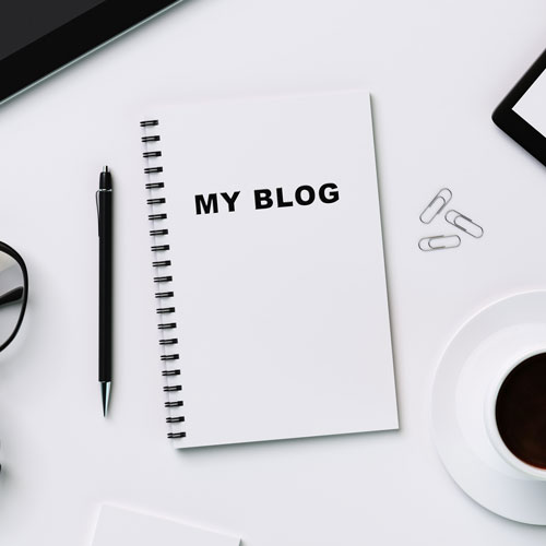 A spiral notebook labelled “MY BLOG” is on a white desk with a pen, spectacles, paper clips, a cup of coffee, and a tablet partially visible—perfect for planning content ideas for a black-owned digital marketing agency in South Africa. - Proudly Developing