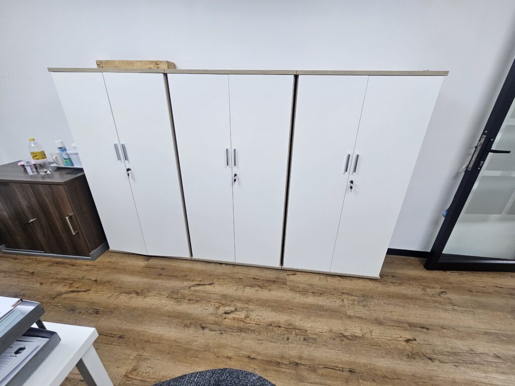 Four white storage cupboards with silver handles stand against a white wall on a wooden floor, next to a brown cupboard and a glass door. - Proudly Developing