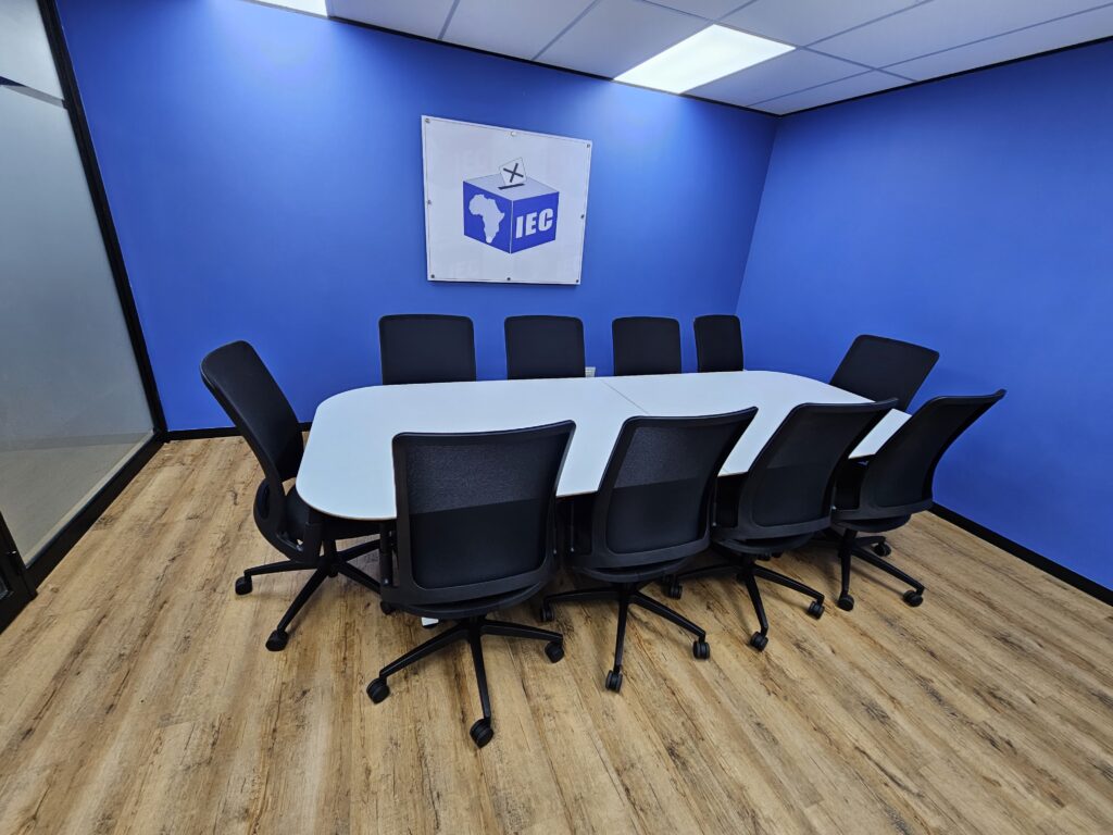A conference room with a long white table, ten black office chairs, wooden flooring, blue walls, and an IEC sign on the wall. - Proudly Developing
