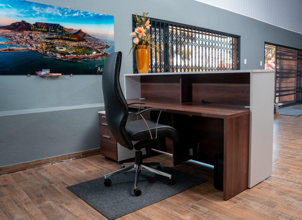 A modern Akandi reception desk with a black office chair, a vase of flowers, and a wall photo of a coastal city in a bright, clean office space. - Proudly Developing
