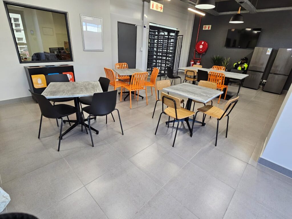 A modern staff room with tables and assorted chairs, vending machines, fridges, a television, a fire hose reel, and a window to an adjoining office. - Proudly Developing